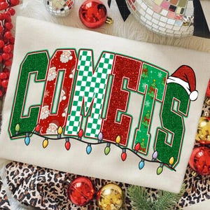 May include: Cream-colored sweatshirt with the word "COMETS" in a festive design. The letters are outlined in green and filled with red glitter, a green and white checkerboard pattern, and a Christmas tree and Santa Claus pattern. A string of colorful lights runs along the bottom.