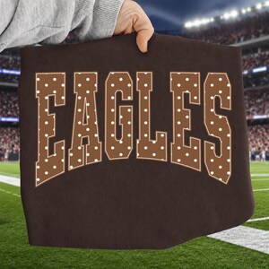 Peut inclure: Un t-shirt marron foncé avec le mot EAGLES écrit en grandes lettres de style bloc. Les lettres sont remplies d'un motif à pois blancs et soulignées d'une fine bordure blanche, disposées en un léger arc au centre du vêtement.