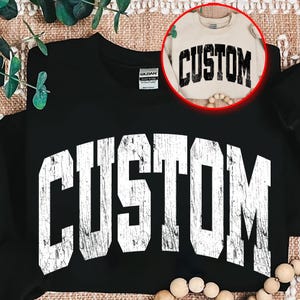 以下が含まれることがあります： 前面に「CUSTOM」の文字が大きく、かすれた白い文字でプリントされた黒いスウェットシャツ。円形のインセットには、「CUSTOM」の文字が入った小さめのクリーム色のスウェットシャツが見えます。スウェットシャツは、テクスチャ加工された表面に表示されています。