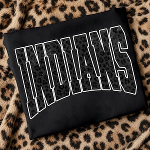 Könnte beinhalten: Ein schwarzes Sweatshirt mit dem Wort INDIANS in großen, bogenförmigen Blockbuchstaben. Der Text hat ein dunkelgraues Leopardenmuster und ist mit einem dicken weißen Rand versehen, der einen starken Kontrast zum schwarzen Stoff bildet.