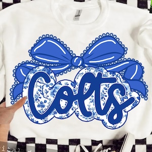 Puede incluir: Sudadera blanca con la palabra "Colts" en diseño floral azul, enmarcada por un lazo azul con borde festoneado. El diseño está centrado en la parte delantera de la prenda.