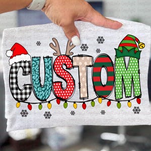 May include: A light gray t-shirt with the word "CUSTOM" in a festive Christmas design. The letters are decorated with patterns like plaid, polka dots, and stripes, and topped with a Santa hat, reindeer antlers, and an elf hat. Colorful Christmas lights are strung below.