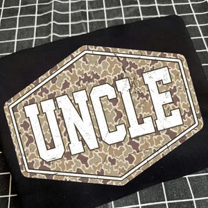 May include: Black t-shirt featuring a distressed, camouflage-patterned hexagon with the word "UNCLE" in bold, white letters. The design has a vintage, worn look. The shirt is folded on a black and white checkered surface.