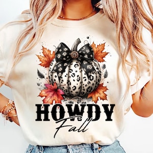 May include: Cream-colored t-shirt featuring a pumpkin design with a black and white leopard print pattern. The pumpkin has a black bow and is surrounded by fall leaves. The text "HOWDY Fall" is printed below the pumpkin.