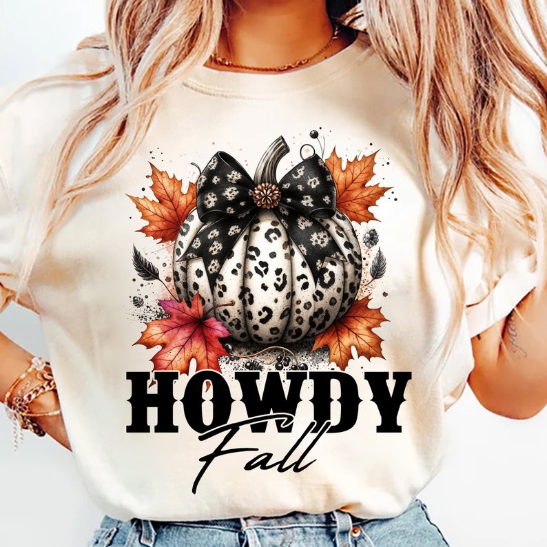 May include: Cream-colored t-shirt featuring a pumpkin design with a black and white leopard print pattern. The pumpkin has a black bow and is surrounded by fall leaves. The text "HOWDY Fall" is printed below the pumpkin.