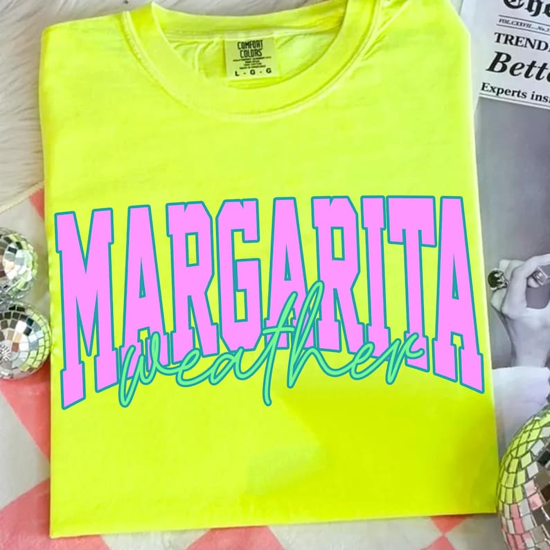 May include: A neon yellow t-shirt with the words "MARGARITA weather" in large pink and teal lettering. The shirt is made of a soft material and has a crew neck. The shirt is a Comfort Colors brand.