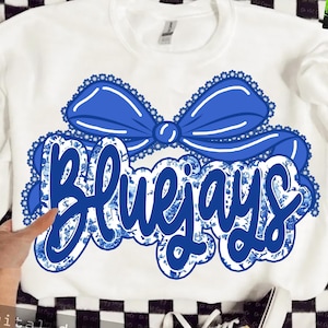 Puede incluir: Sudadera blanca con un gran lazo azul y la palabra "Bluejays" en una fuente decorativa. El texto tiene un patrón floral azul. El lazo está delineado con un ribete de encaje festoneado.
