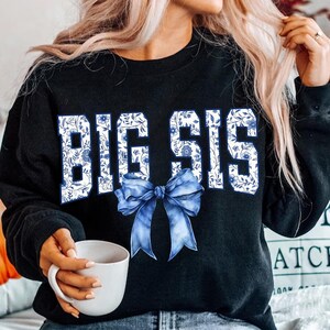 May include: Black sweatshirt with the words "Big Sis" in white letters with a blue and white floral pattern. A blue bow is centered below the text.