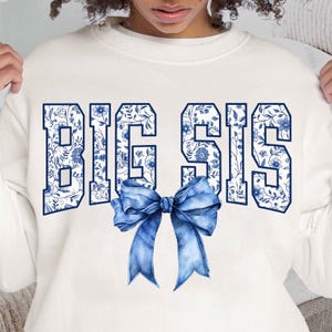 May include: White sweatshirt with the words "BIG SIS" in blue letters with a blue and white floral pattern. A blue bow is centered below the text.