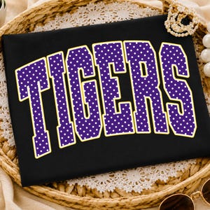 Puede incluir: Una camiseta negra con la palabra TIGERS en una fuente grande y arqueada de estilo universitario. Las letras son de color morado con un patrón de puntos blancos y un borde amarillo fino, creando un diseño gráfico llamativo en la prenda.