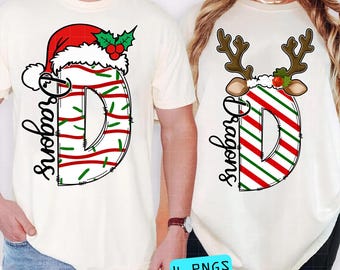Puede incluir: Dos camisetas color crema con un diseño de "D" con temática navideña. Una presenta un gorro de Papá Noel, la otra cuernos de reno. La "D" está a rayas rojas y verdes con la palabra "Dragons" en escritura negra. La parte inferior de la imagen tiene el texto "4 PNGS".