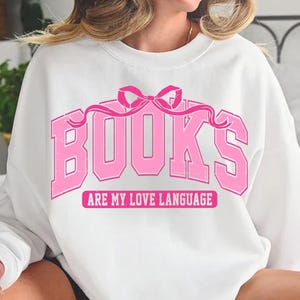 Pu&ograve; includere: Felpa bianca con una grafica rosa che dice "BOOKS ARE MY LOVE LANGUAGE" con un fiocco rosa sopra il testo.