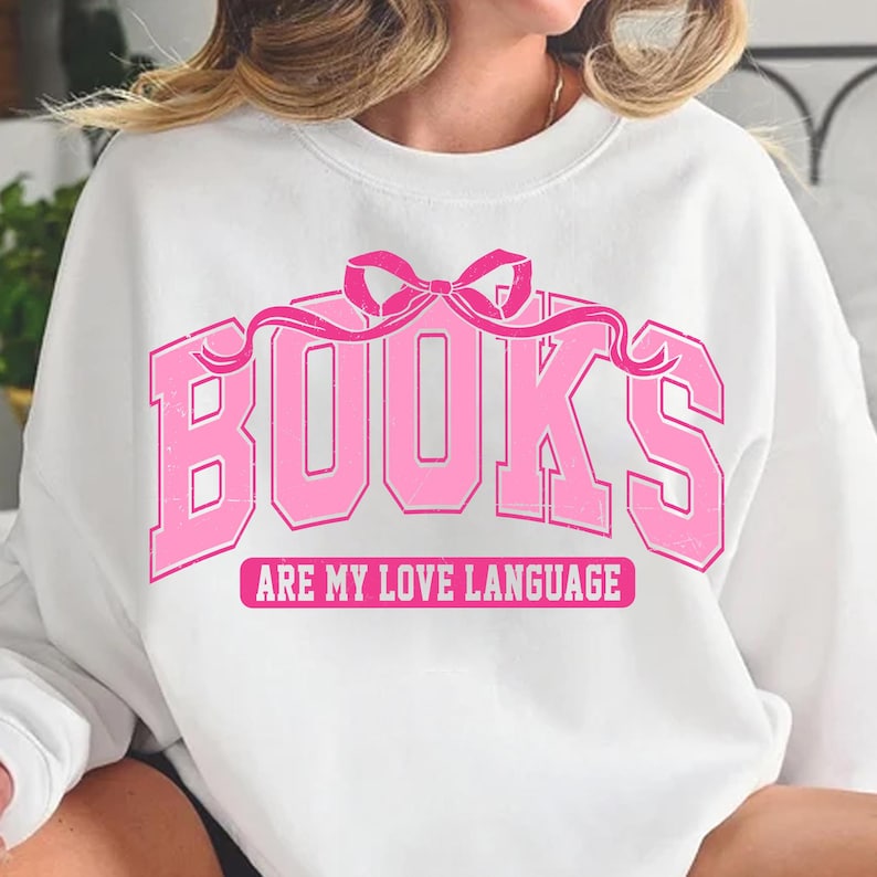 Pu&ograve; includere: Felpa bianca con una grafica rosa che dice "BOOKS ARE MY LOVE LANGUAGE" con un fiocco rosa sopra il testo.