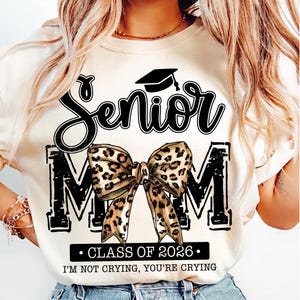 May include: Cream-colored t-shirt with a black graphic that says "Senior Mom" with a graduation cap and a leopard print bow. The text "Class of 2026" and "I'M NOT CRYING, YOU'RE CRYING" are also printed on the shirt.