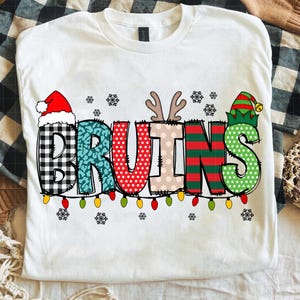 以下が含まれることがあります： 「BRUINS」の文字がデザインされた白いTシャツ。文字はチェック柄、水玉模様、ストライプなどのパターンで装飾され、サンタ帽、トナカイの角、エルフの帽子が飾られています。文字の下には電飾が走っています。