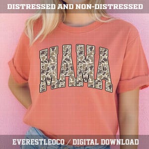 May include: A coral-colored sweatshirt with the word "MAMA" in large, block letters across the front. The letters are filled with a camouflage pattern in shades of brown and tan. The sleeves are rolled up. The text "DISTRESSED AND NON-DISTRESSED" is at the top.