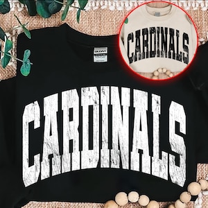 May include: Black sweatshirt with the word "CARDINALS" in large, distressed white lettering across the front. A smaller, tan sweatshirt with the same design is visible in a circular inset. The design is a vintage style.