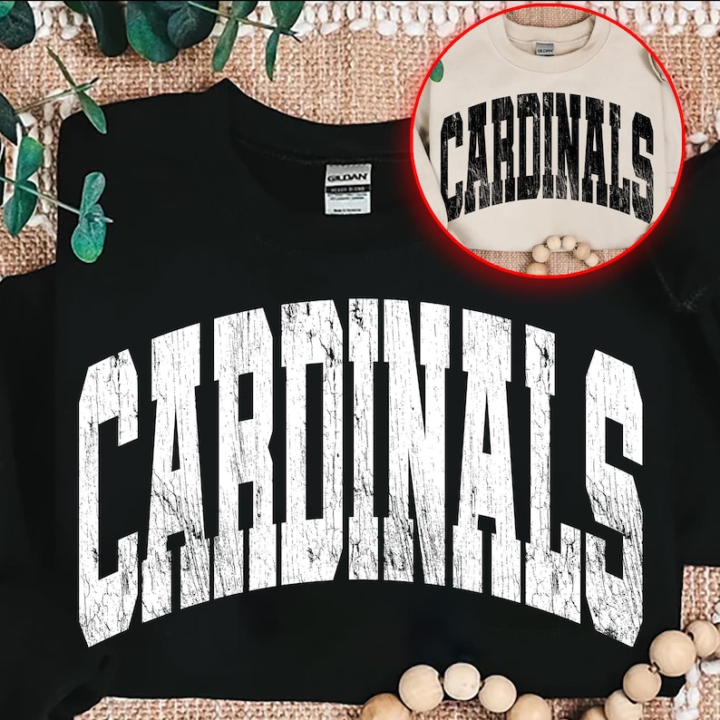 May include: Black sweatshirt with the word "CARDINALS" in large, distressed white lettering across the front. A smaller, tan sweatshirt with the same design is visible in a circular inset. The design is a vintage style.