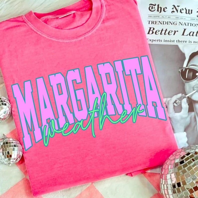 May include: Bright pink t-shirt with the words "MARGARITA weather" in large, blocky, pink letters with a teal outline. The word "weather" is in a cursive, green font. The shirt is on a pink and white checkered surface.