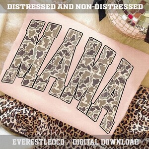 May include: A light pink t-shirt with the word "MAMA" in large, distressed camouflage letters. The shirt is displayed on a soft, textured surface with a leopard print fabric visible below. The text "DISTRESSED AND NON-DISTRESSED" is at the top.