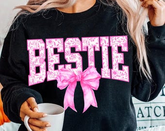 Op de afbeelding: Zwart sweatshirt met het woord "BESTIE" in roze bloemenletters en een roze strik. Een casual stijl, ideaal als cadeau of voor dagelijks gebruik.