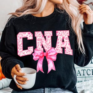 May include: Black sweatshirt with a pink floral design spelling out "CNA" with a pink bow in the center.