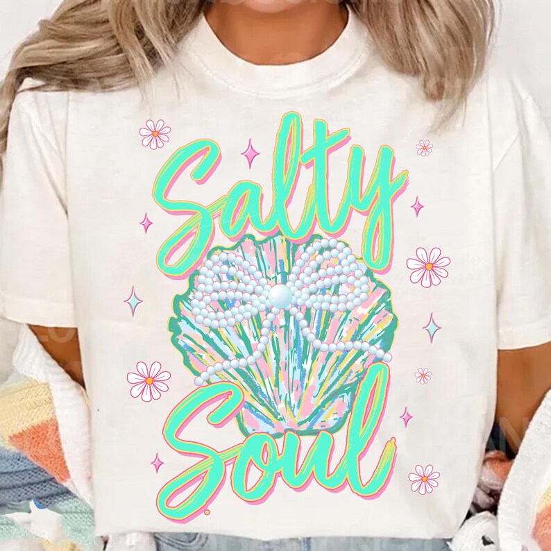 May include: White t-shirt featuring a green and pink graphic design of a seashell with pearls and the text "Salty Soul".