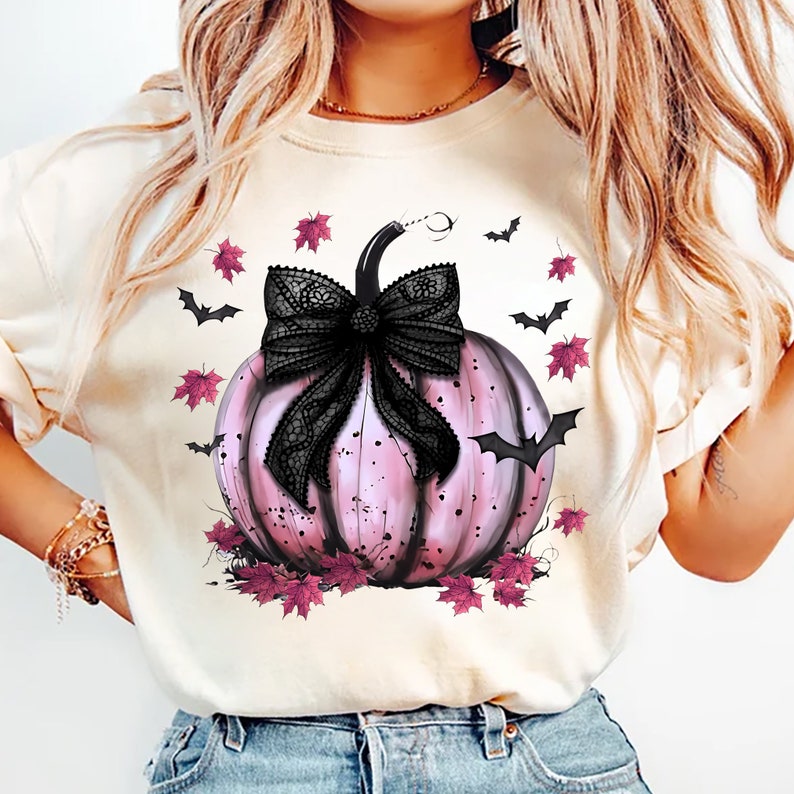 May include: A cream-coloured sweatshirt featuring a pink pumpkin with a black lace bow. The pumpkin is surrounded by black bats and pink leaves, creating a Halloween theme. The design is a graphic print.