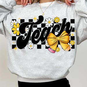 May include: Gray sweatshirt with a black and white checkered background. The word "Teach" is written in a retro font with a yellow bow and pencil. The design is surrounded by yellow and white flowers.
