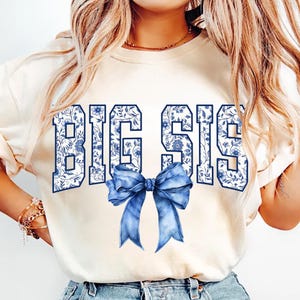 May include: A white t-shirt with the words "BIG SIS" printed in blue with a blue floral pattern. A blue bow is centered below the text.