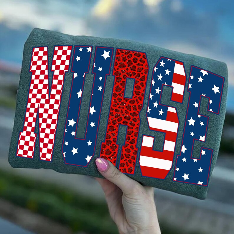 May include: A gray t-shirt with the word "NURSE" in a patriotic design. The letters feature red and white checkered patterns, blue with white stars, and red leopard print. The shirt is held up against a blurred blue sky.