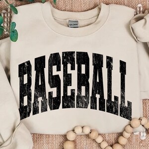 May include: Cream-colored sweatshirt with the word "BASEBALL" printed in large, distressed black letters across the front. The sweatshirt has a crew neck and long sleeves. The Gildan brand tag is visible.