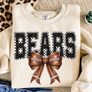 May include: A cream-colored crewneck sweatshirt featuring the word BEARS in large, black, leopard-print block letters with a white and black checkered border. Below the text is a brown football-textured bow with white laces and silver accents.