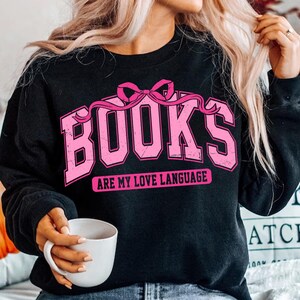 Pu&ograve; includere: Felpa nera con una grafica rosa che dice "BOOKS ARE MY LOVE LANGUAGE" con un fiocco rosa sopra la parola "BOOKS".