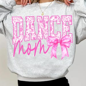 May include: A light gray sweatshirt with the words "DANCE Mom" in pink. The word "DANCE" is filled with a floral pattern. A large pink bow is on the right side of the text.