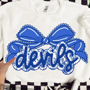 May include: White sweatshirt featuring a large blue bow with a scalloped edge and the word "devils" in a blue floral design. The design is centered on the front of the shirt. The sweatshirt is a Gildan brand.