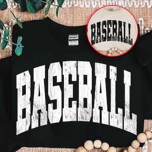 May include: Black sweatshirt with the word "BASEBALL" in large, distressed white letters across the front. A smaller, circular image shows a tan shirt with the word "BASEBALL" in black. The sweatshirt is folded on a textured surface.