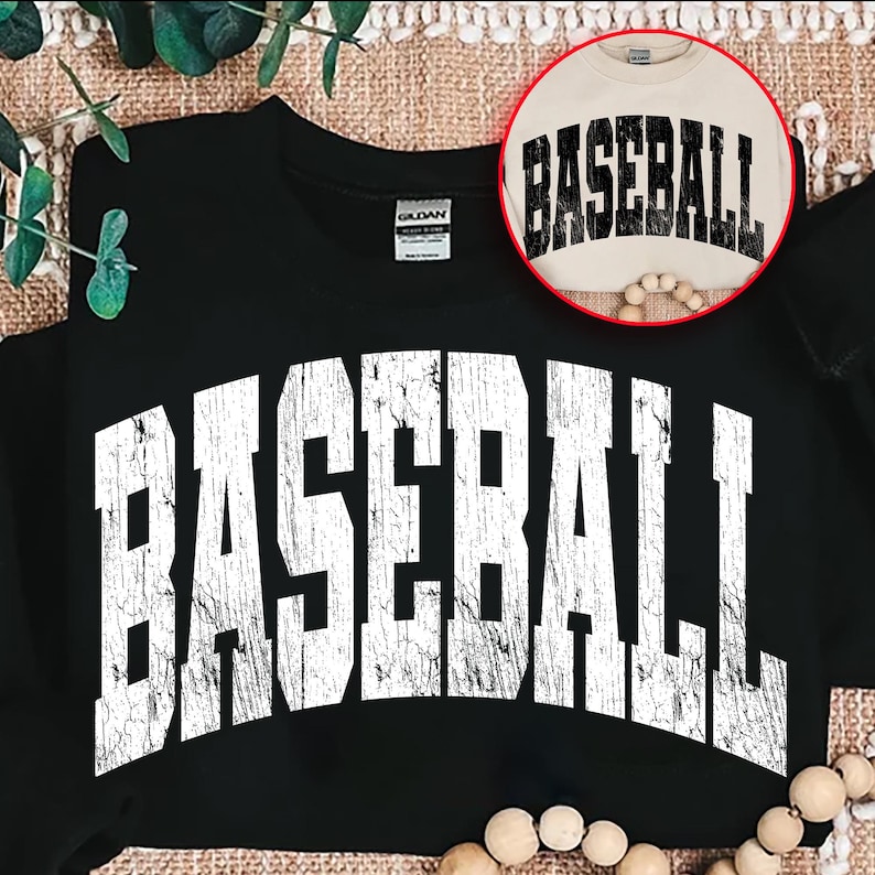 May include: Black sweatshirt with the word "BASEBALL" in large, distressed white letters across the front. A smaller, circular image shows a tan shirt with the word "BASEBALL" in black. The sweatshirt is folded on a textured surface.
