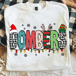 May include: White t-shirt with the word "BOMBERS" in a festive design. The letters are decorated with patterns like buffalo check, polka dots, and stripes, and topped with a Santa hat, antlers, and an elf hat. A string of colorful lights runs below the word.