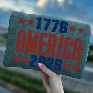Op de afbeelding: Een groen t-shirt met de tekst "1776 AMERICA 2026" in verweerde rode en blauwe letters. Het ontwerp bevat rode sterren en lijnen, wat een patriottisch thema suggereert. Het t-shirt wordt tegen een vage achtergrond gehouden.