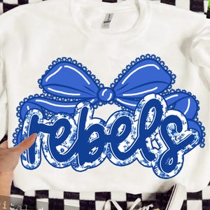 May include: White sweatshirt with a blue bow and the word "rebels" in a blue floral design. The word "rebels" is outlined in blue and filled with a blue floral pattern. The bow is a solid blue color with a white lace trim.
