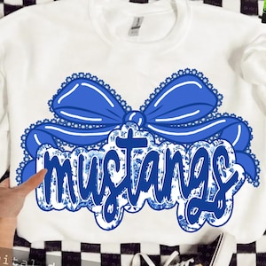 May include: White sweatshirt featuring a large blue bow with a scalloped edge and the word "mustangs" in a floral pattern. The text is in a bold, stylized font. The design is centered on the front of the shirt.