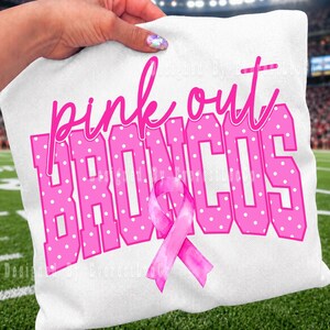 Puede incluir: Una camiseta blanca con el texto rosa "pink out" en letra cursiva sobre la palabra "BRONCOS" en letras grandes, rosas y en bloque con lunares blancos. Un lazo rosa de concienciación está colocado sobre el centro de la palabra "BRONCOS".