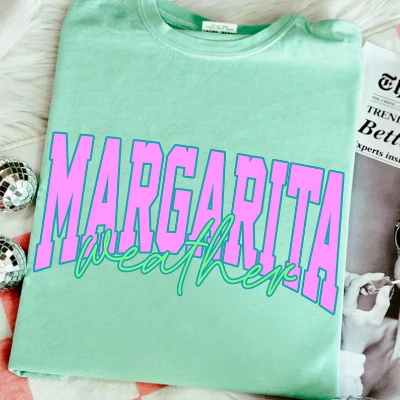 May include: A light green t-shirt with the words "MARGARITA weather" in large pink block letters with a green outline. The shirt is folded and resting on a white surface. The shirt is a crew neck.
