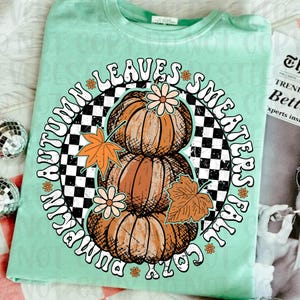 May include: A light green t-shirt featuring a graphic design with three stacked pumpkins, fall leaves, and the text "Autumn Leaves, Sweater Weather, Cozy Fall, Pumpkin Spice". The design is set against a black and white checkered background.
