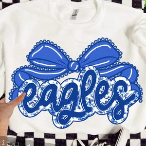 May include: White sweatshirt with a large blue bow and the word "eagles" in a blue floral design. The bow has a scalloped edge. The word "eagles" is in a script font.