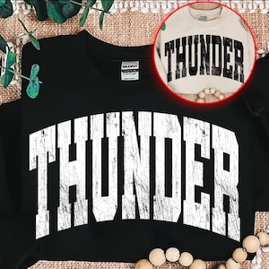 Może przedstawiać: Czarna bluza z napisem "THUNDER" w dużych, postarzanych białych literach z przodu. Drugi, beżowy sweter z tym samym wzorem jest pokazany w okrągłej wstawce. Projekt ma wygląd vintage.