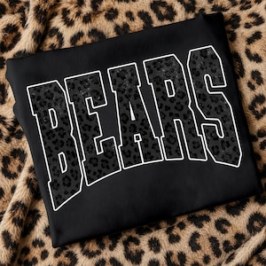 May include: A black sweatshirt featuring the word BEARS in large, block-style varsity lettering. The letters are filled with a dark gray and black leopard print pattern and outlined in a crisp white border, creating a bold, high-contrast design.