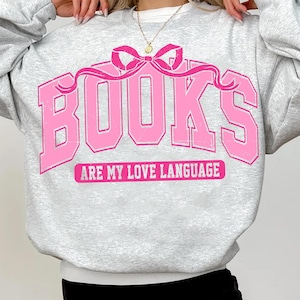 Pu&ograve; includere: Una felpa grigia con una grafica rosa che dice "BOOKS ARE MY LOVE LANGUAGE" con un fiocco rosa sopra la parola "BOOKS".
