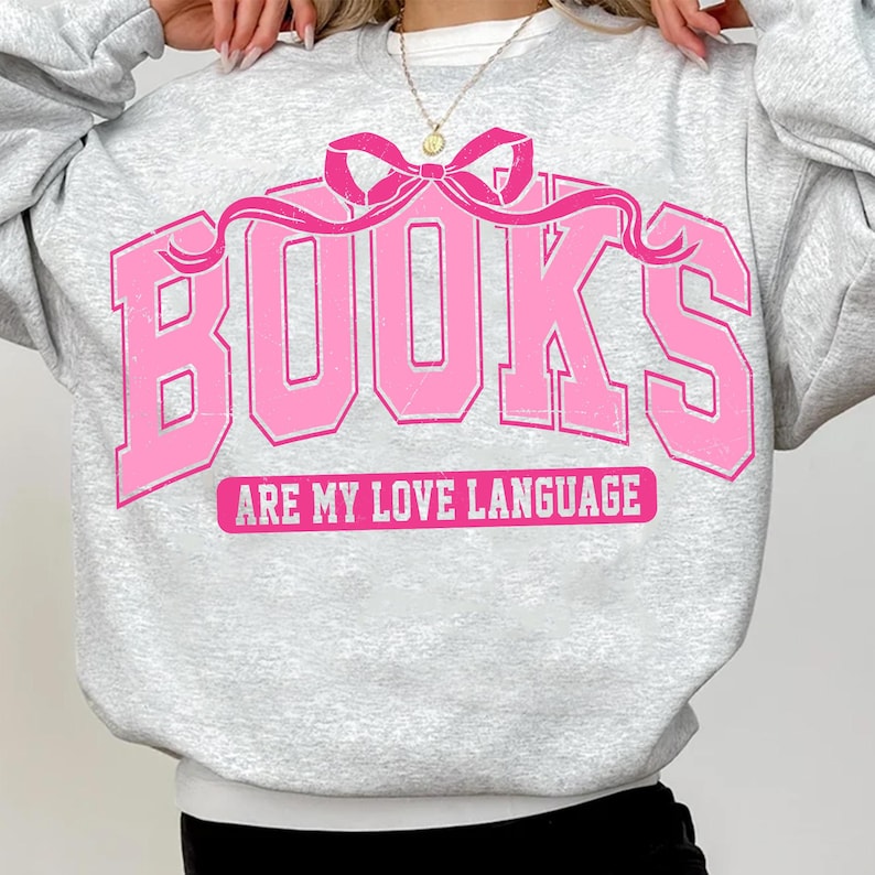 Pu&ograve; includere: Una felpa grigia con una grafica rosa che dice "BOOKS ARE MY LOVE LANGUAGE" con un fiocco rosa sopra la parola "BOOKS".